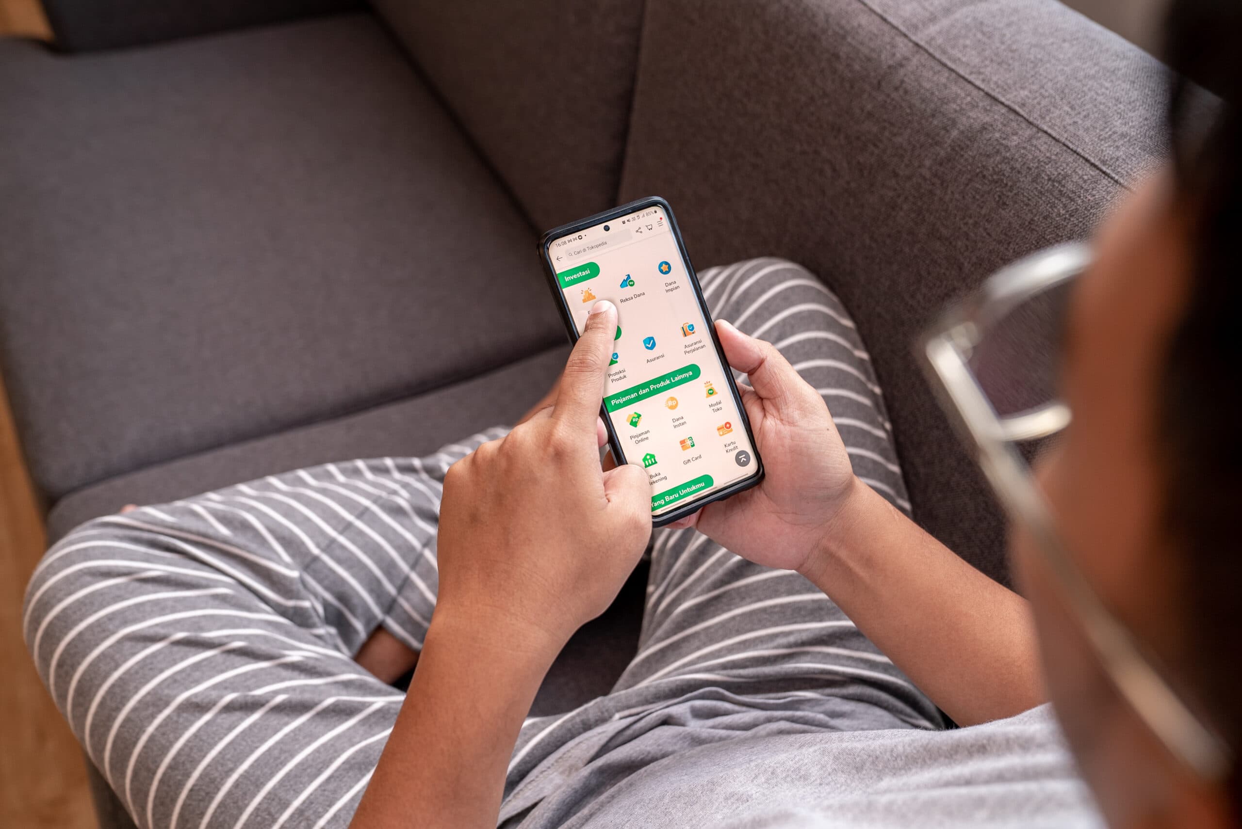 A person sits on a couch while using the Tokopedia app on their phone.